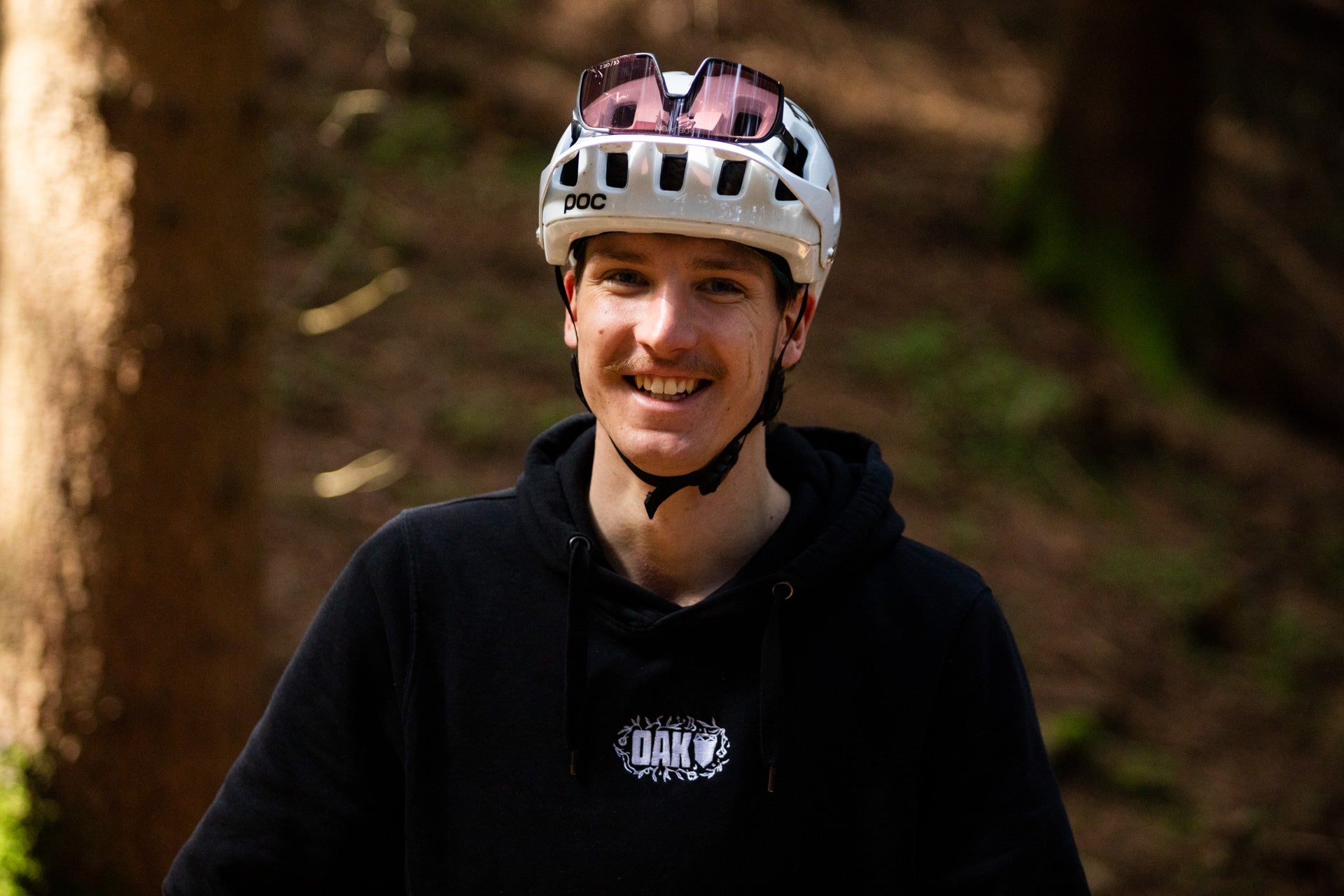 Smiling young man wearing white cycling helmet with pink sunglasses and black hoodie in a forest setting