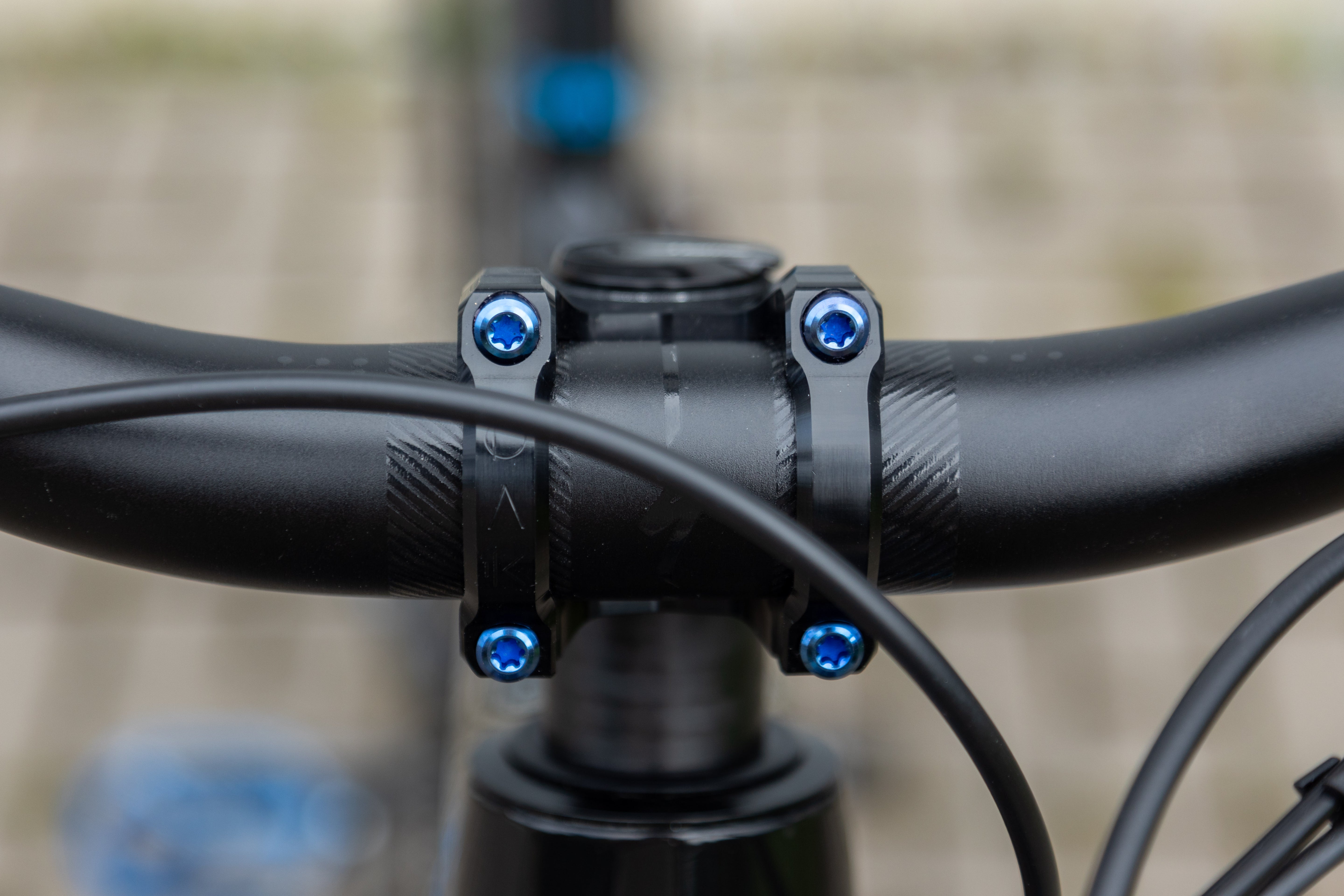 Black mountain bike handlebar with four blue screws and cable, blurred ground and blue pedal in background