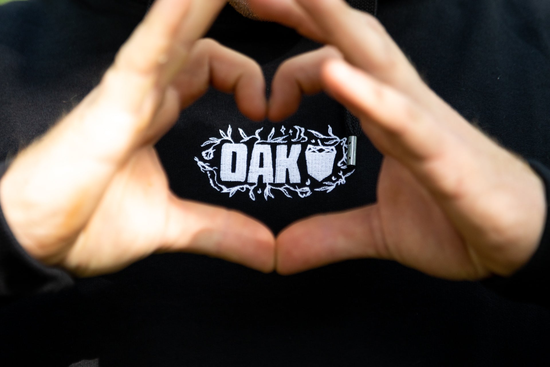 Close-up of hands forming a heart shape over black OAK garment with white acorn embroidery