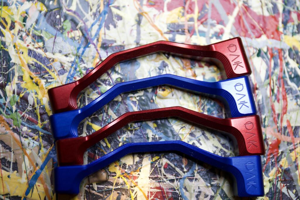 Four metallic mountain bike components in red and blue, engraved with 'OAK', on a paint-splattered background.