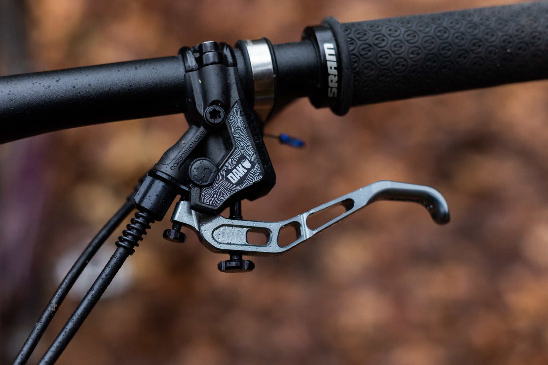 Close-up of OAK mountain bike brake lever with CORBOTECTURE branding and SRAM grip on wet handlebar.