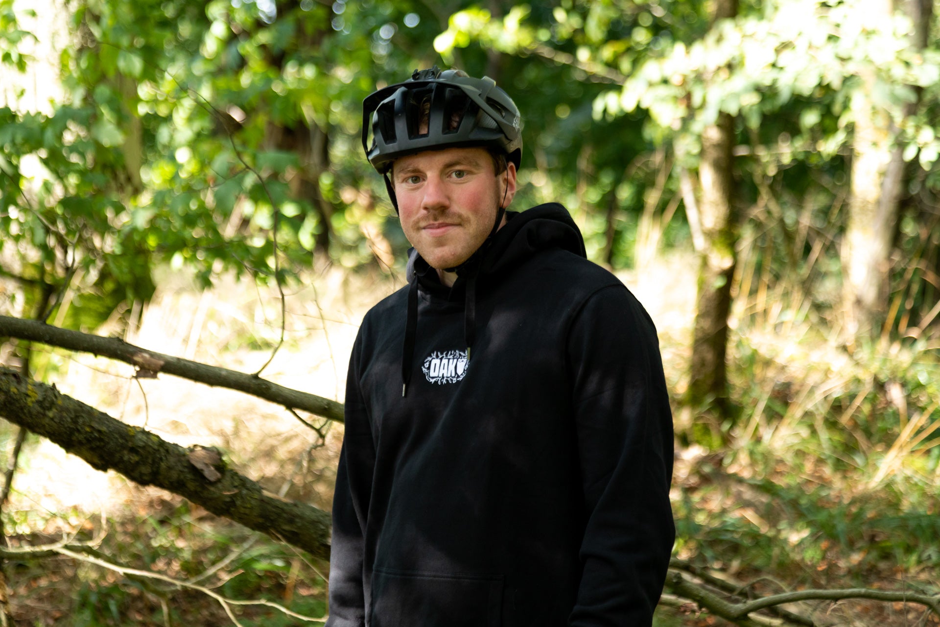 Young man wearing a black bicycle helmet and hoodie in a sunlit green forest.