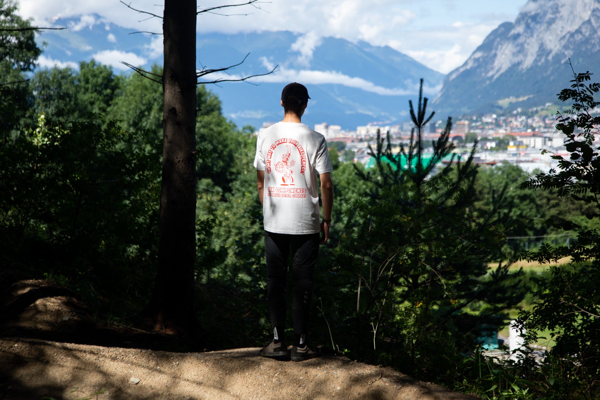 Person on forest path wearing white T-shirt with red OAK Components logo, city and mountains in background