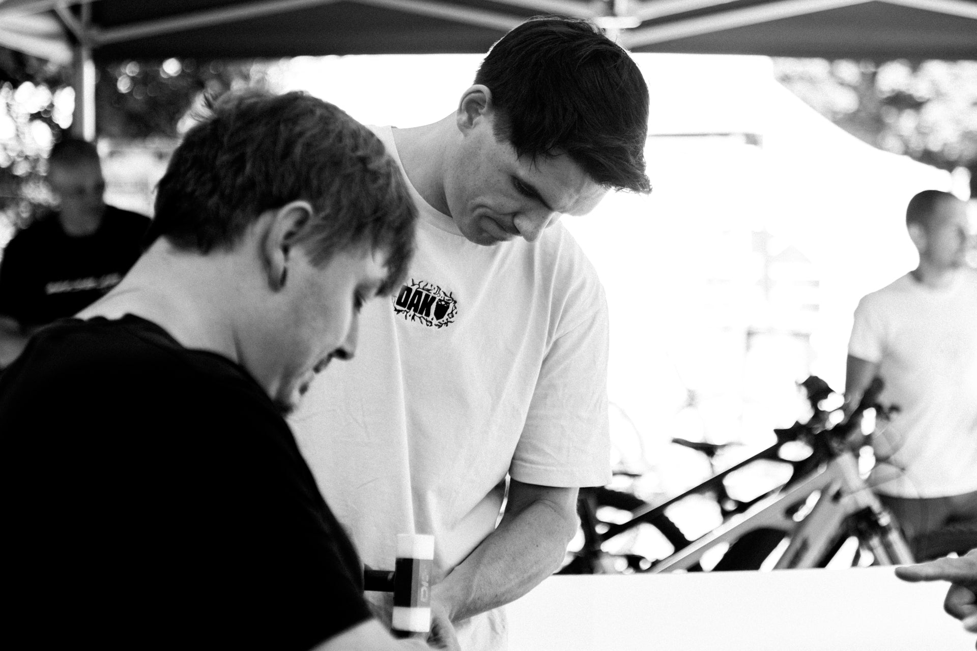 Two young men under a black canopy, one wearing a white t-shirt with text, at an outdoor cycling event.
